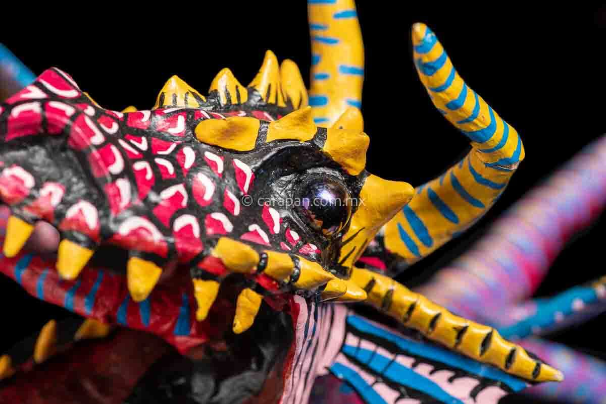 Mexican Paper Mache Alebrije  Dragon  style with fierce expression, two horns paionted in yellow with blue strokes, marble eyes, open mouth showing tootn, long red tongue with blue strokes, wings decorated in blue and pink tones on the outside and blue and white on the inside. Long tail decorated in red with light blue patches with yellow center. HAnds and legs decorated in blue with yellow dots. 