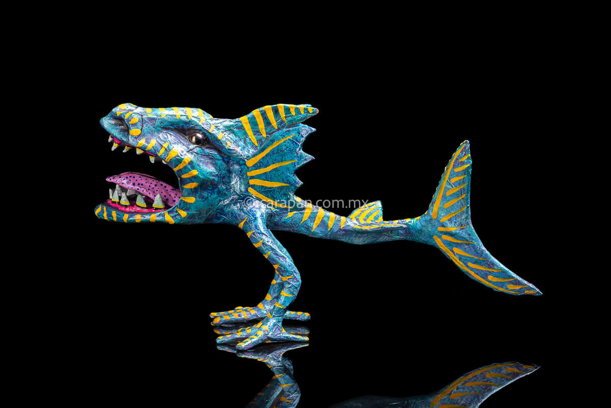 Paper Mache Alebrije inspired by sharks in teal and turquoise tones with yellow strokes, fierce open mouth showing tooth and pink tongue with blue dots. Standing on two legs  left