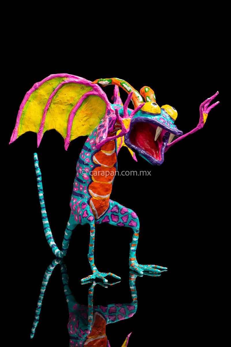 Paper Mache Alebrije Dragon Style Standing with hands up and mouth open showing 2 fangs popping yellow eyes and two orange horns. His body is painted in turquoise with pink dots and blue stripes and his wings are yellow outlined in pink 