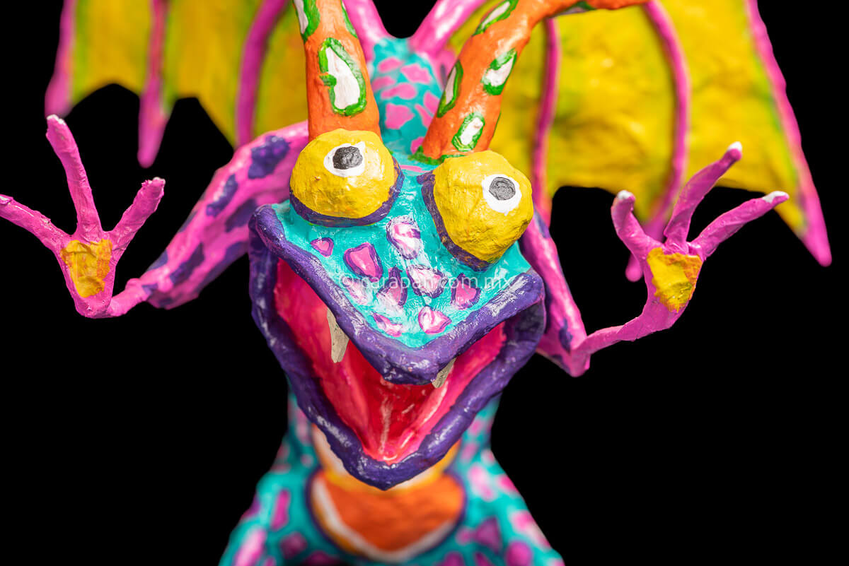 User  Please create a selling description for Paper Mache Alebrije Dragon Style Standing with hands up and mouth open showing 2 fangs popping yellow eyes and two orange horns. His body is painted in turquoise with pink dots and blue stripes and his wings are yellow outlined in pink