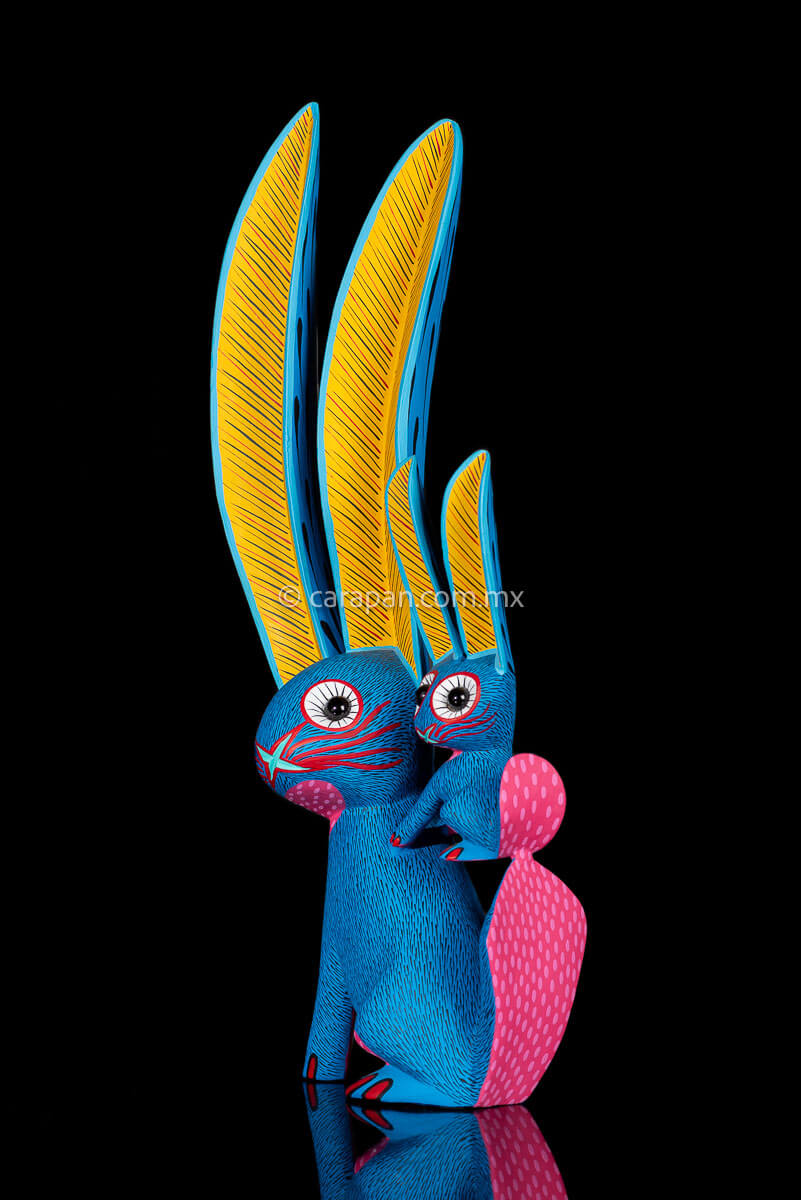 Rabbit with a bunny on its back wooden sculpture the body is painted in blue. the chest in pink and the ears in contrasting yellow the whole piece is decorated with fine strokes in black
