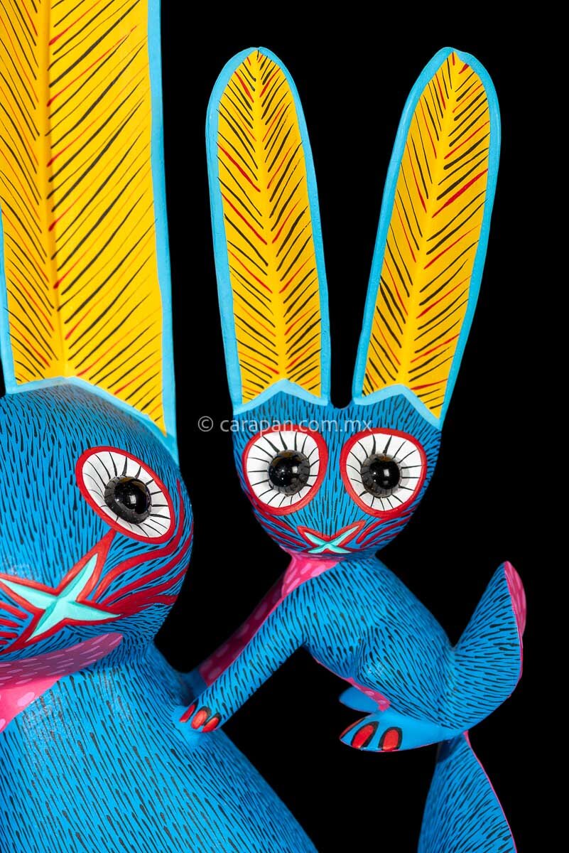 Rabbit with a bunny on its back wooden sculpture the body is painted in blue. the chest in pink and the ears in contrasting yellow the whole piece is decorated with fine strokes in black