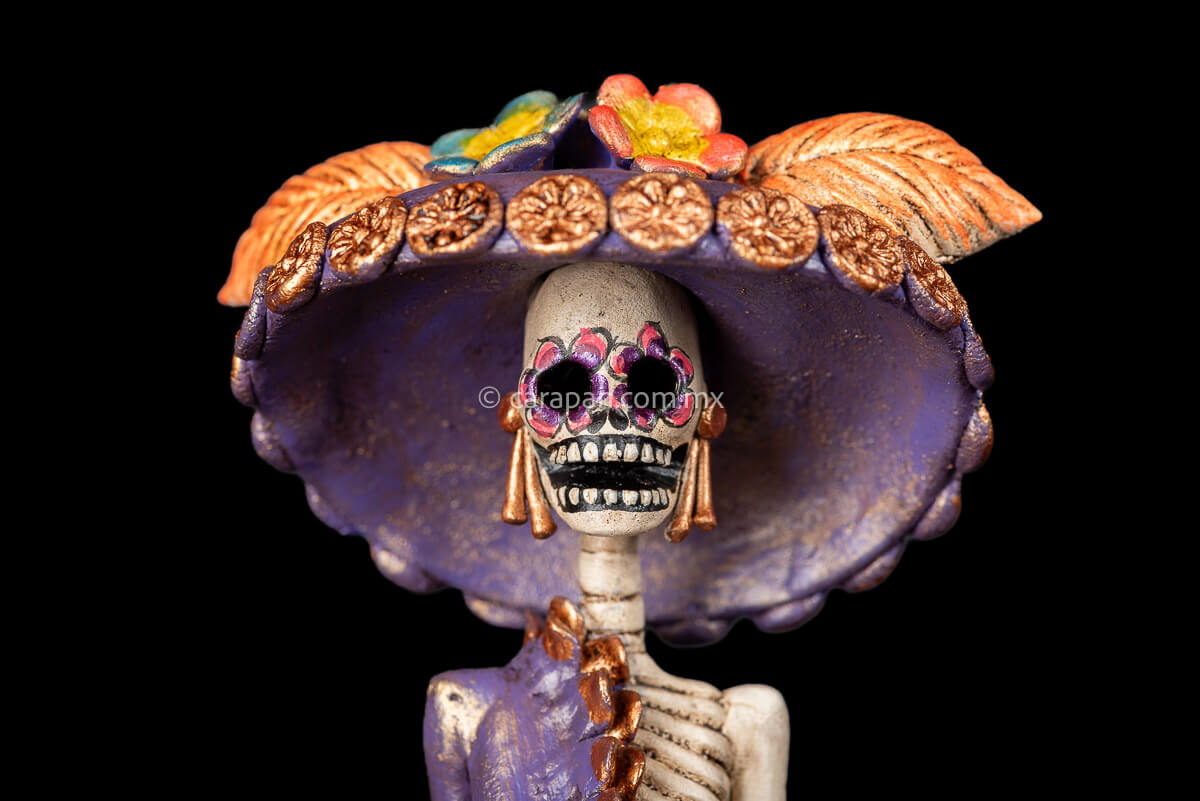 Clay Sculpture of Mexican Catrina, Day of the dead icon inspired by Jose guadalupe posada's work wearing a purple dress with one sleeve, decorated with flowers at the bottom and wearing a traditional hat