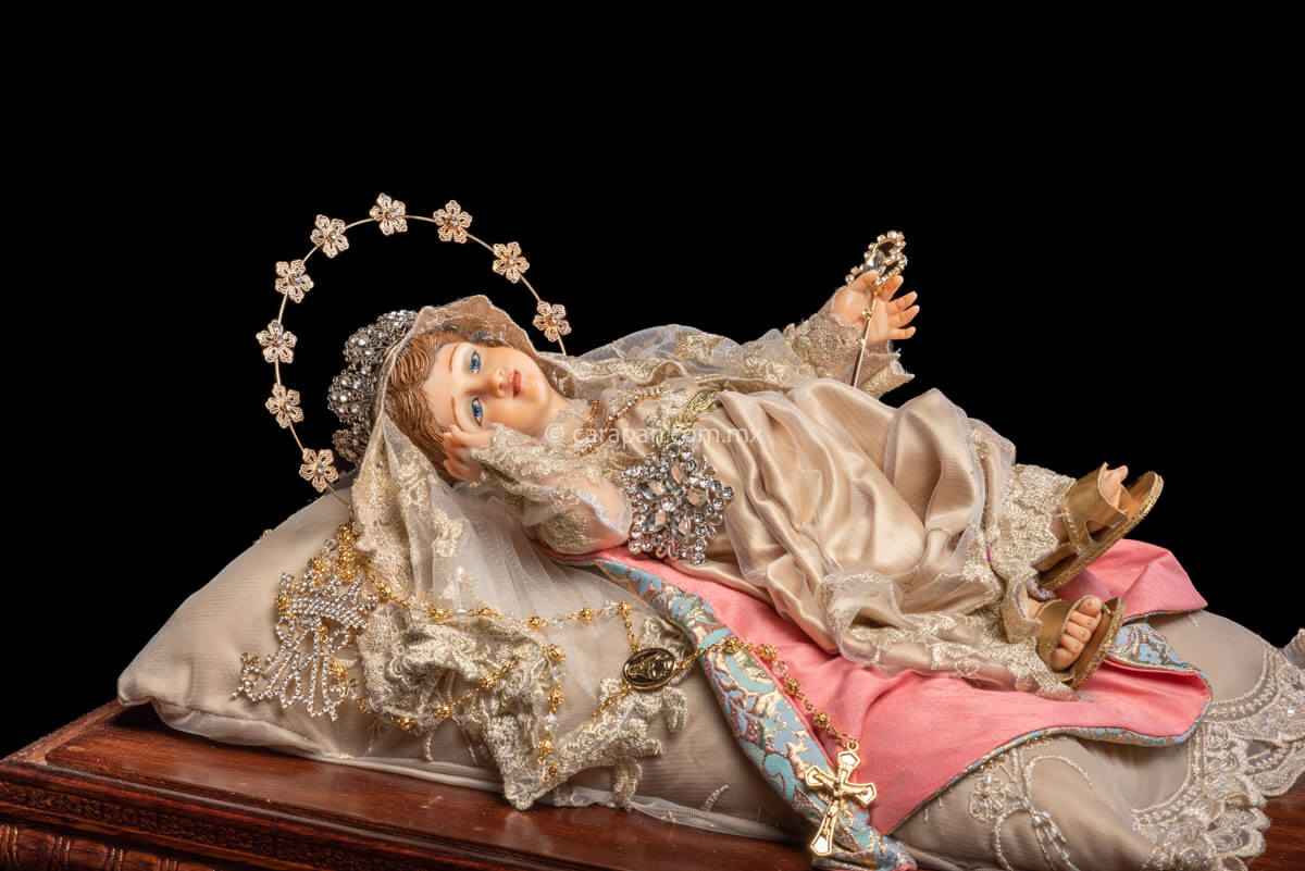 Holy Infant Mary laying position Wax sculpture wearing a silver metal crown with cristals. She holds a scepter in her left hand while her face rests near her right hand.  Her dress is birght gray and has golden touches . A rosary is part of the ensemble. A golden metal halo with flowers and cristal center resembles her aura. 