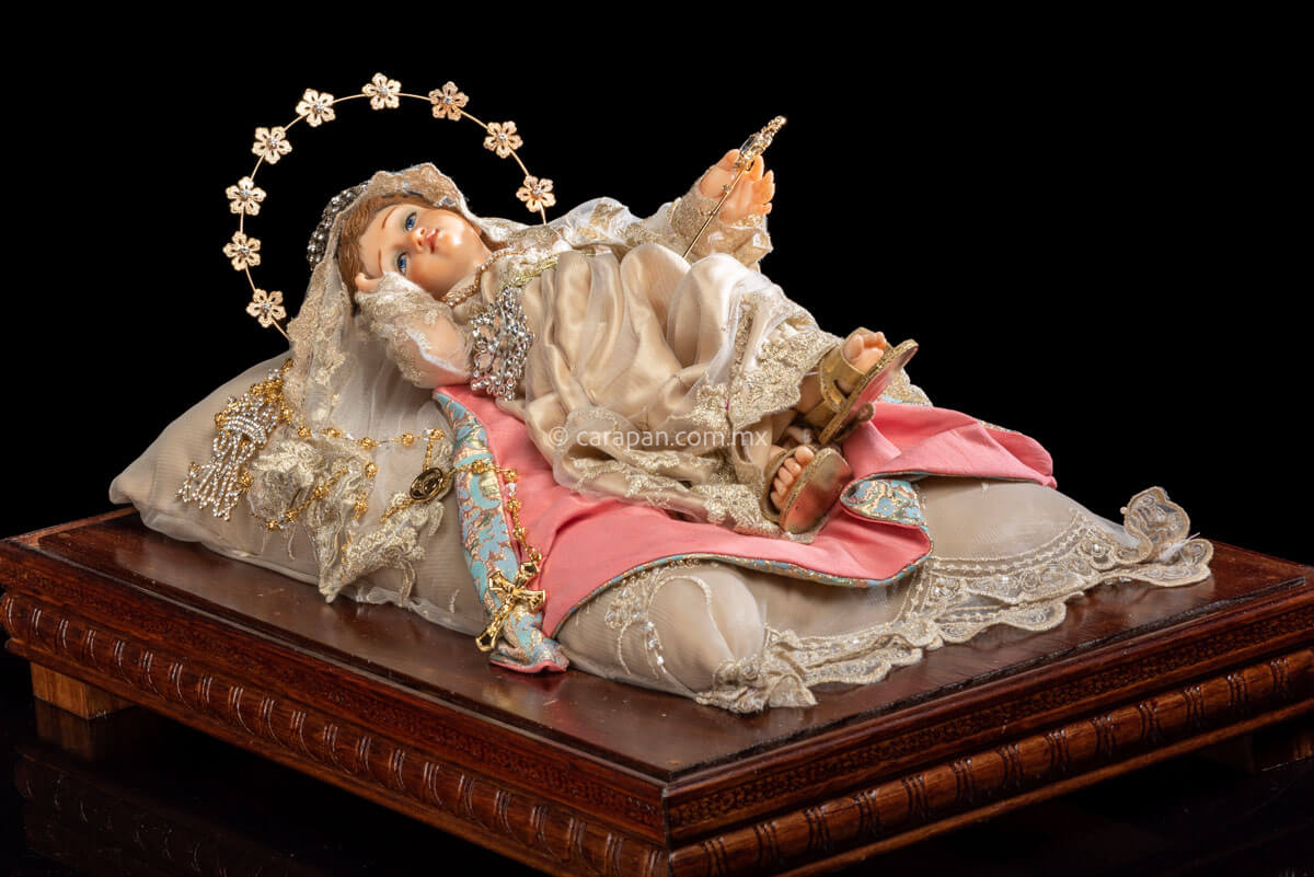     Holy Infant Mary laying position Wax sculpture wearing a silver metal crown with cristals. She holds a scepter in her left hand while her face rests near her right hand.  Her dress is birght gray and has golden touches . A rosary is part of the ensemble. A golden metal halo with flowers and cristal center resembles her aura. 